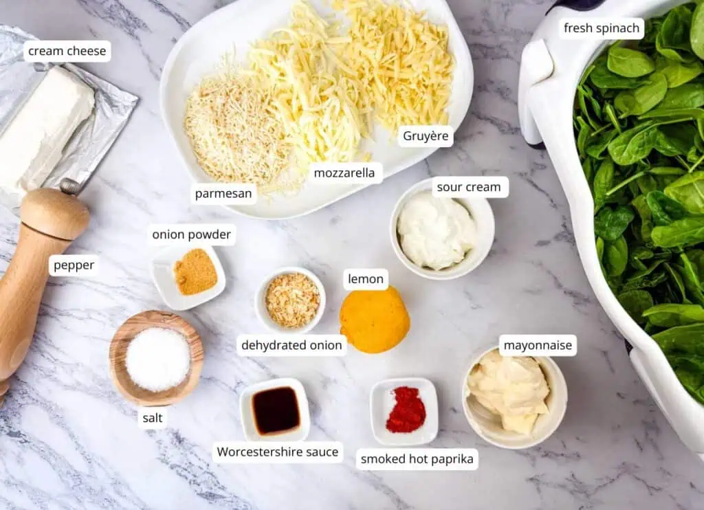 Ingredients for a spinach dip, including cream cheese, pepper, salt, lemon, grated cheese, sour cream, mayonnaise, spices, Worcestershire sauce, and fresh spinach, arranged on a marble surface.