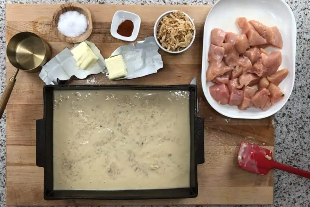 Ingredients for a casserole are arranged on a wooden board, including cubed raw chicken, butter, spices, crispy onions, and a rectangular pan with a creamy sauce.