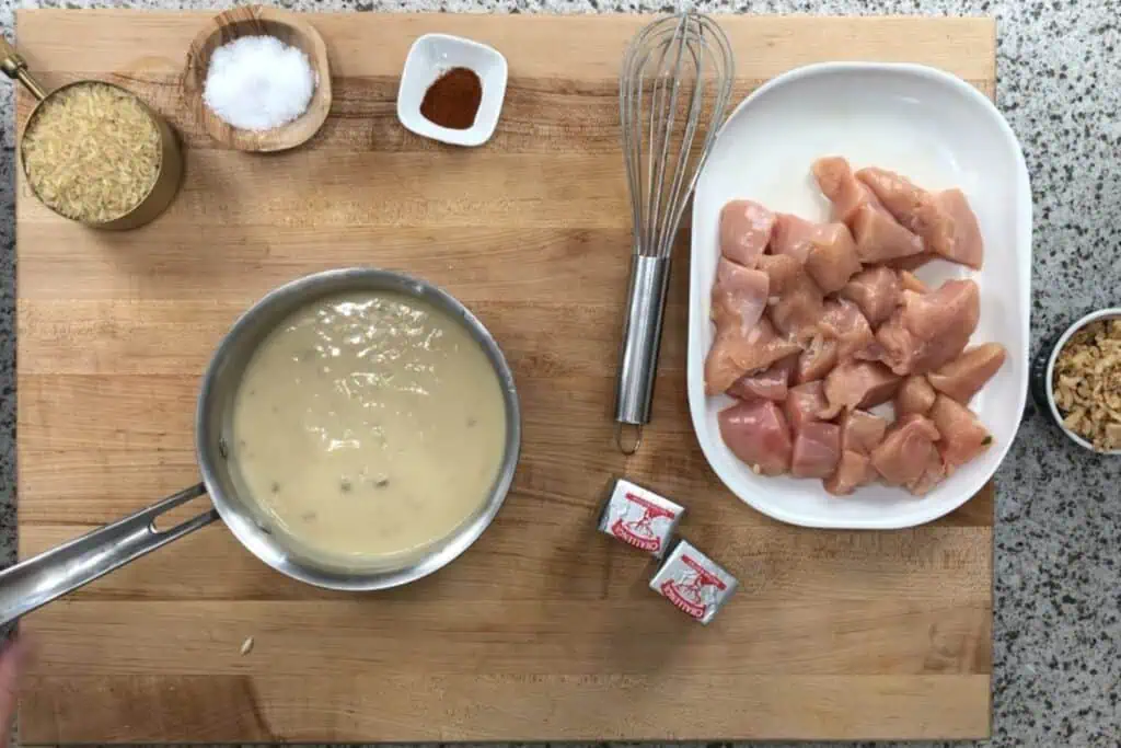 A wooden cutting board with raw diced chicken, two bouillon cubes, crispy fried onions, uncooked rice, salt, paprika, a whisk, and a pot of creamy sauce.