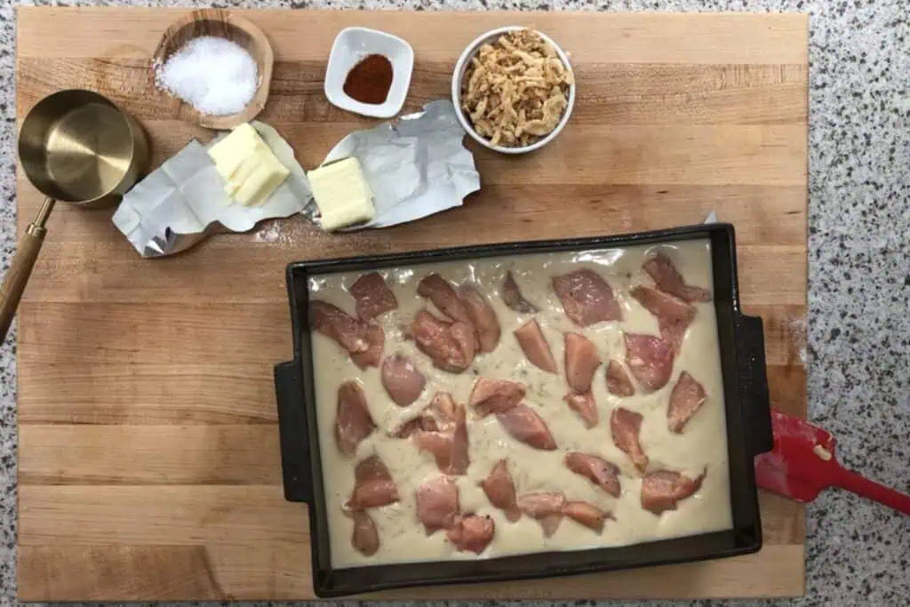 A baking pan with uncooked chicken pieces and creamy sauce sits on a wooden board, surrounded by butter, salt, paprika, fried onions, and a measuring cup.