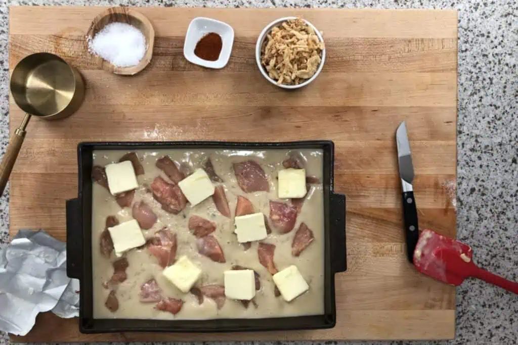 A baking pan with raw chicken pieces, sauce, and butter pats on a wooden board, surrounded by salt, paprika, fried onions, a measuring cup, butter wrapper, and a spatula.