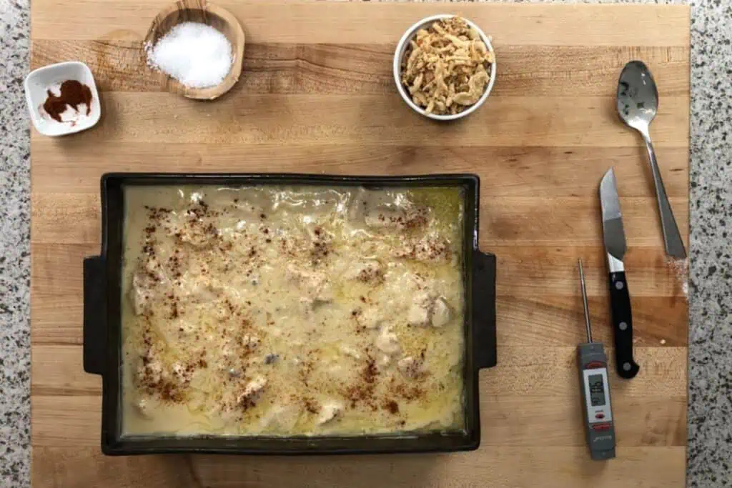 Rectangular baking dish filled with a creamy, baked casserole sits on a wooden surface, surrounded by a bowl of salt, a bowl of fried onions, a spoon, a thermometer, and a paring knife.