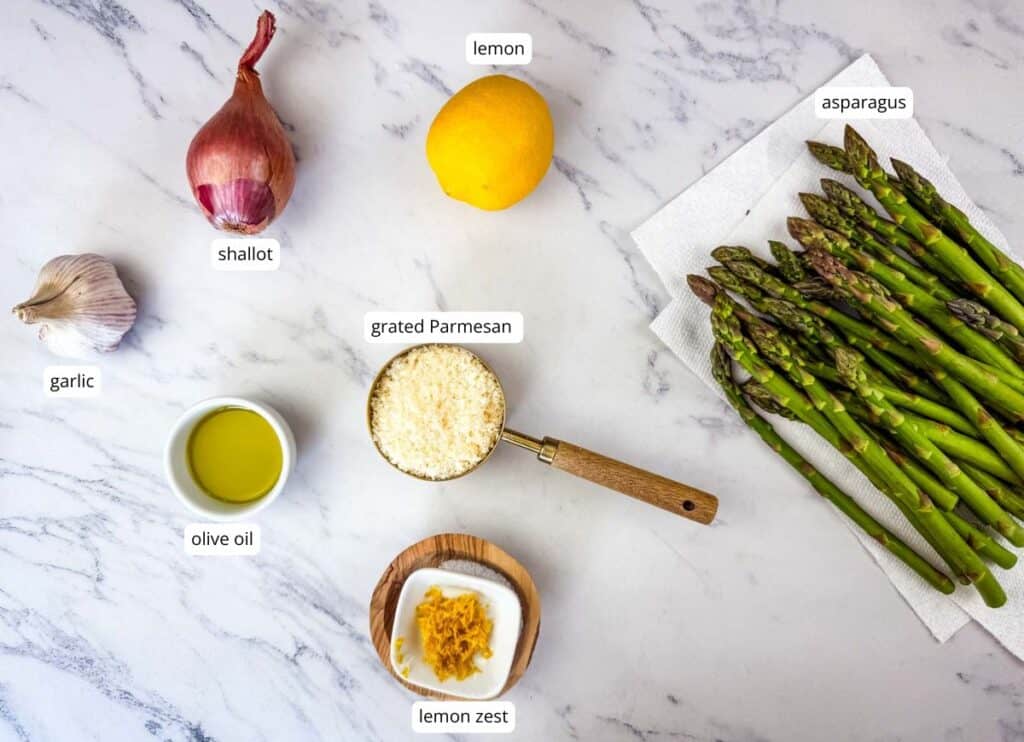 Asparagus, lemon, shallot, garlic, olive oil, grated Parmesan, and lemon zest are arranged on a marble surface; the asparagus rests on a paper towel.