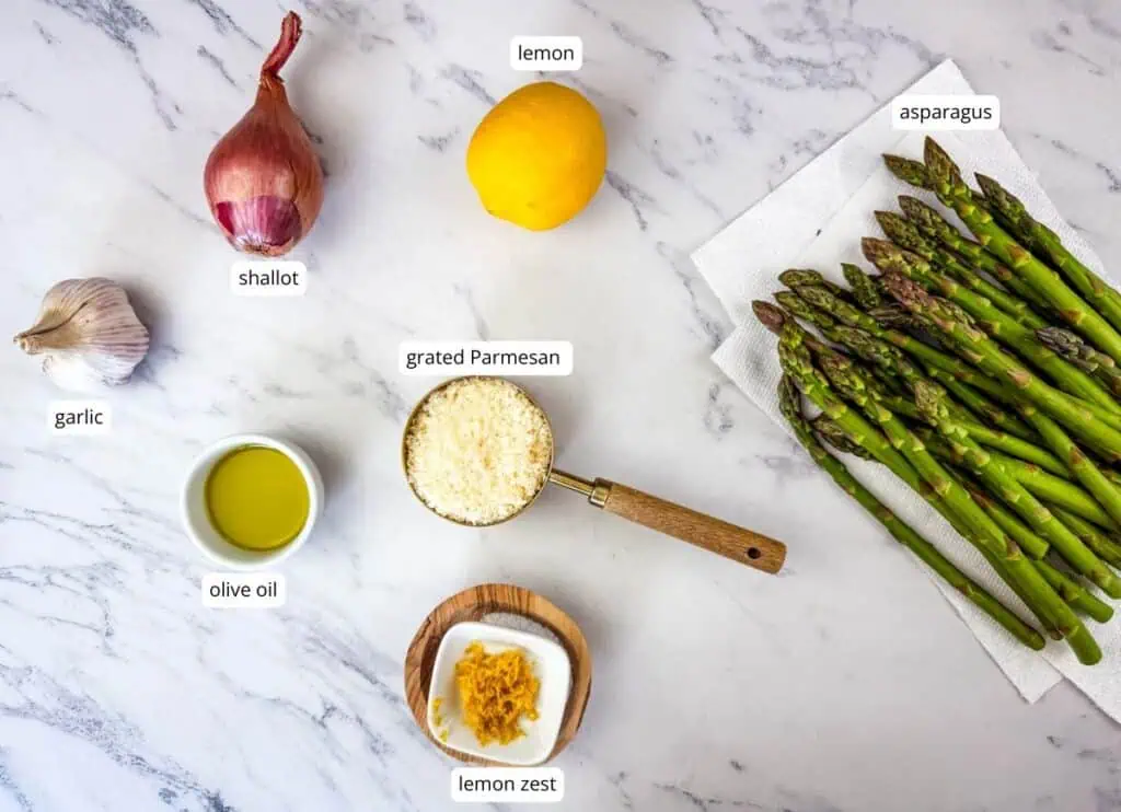 Asparagus, lemon, shallot, garlic, olive oil, grated Parmesan, and lemon zest are arranged on a marble surface; the asparagus rests on a paper towel.