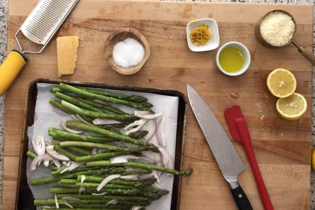 A cutting board with asparagus and sliced onions on a tray, a chef&rsquo;s knife, lemon, grated cheese, olive oil, salt, lemon zest, a cheese grater, and a spatula arranged nearby.