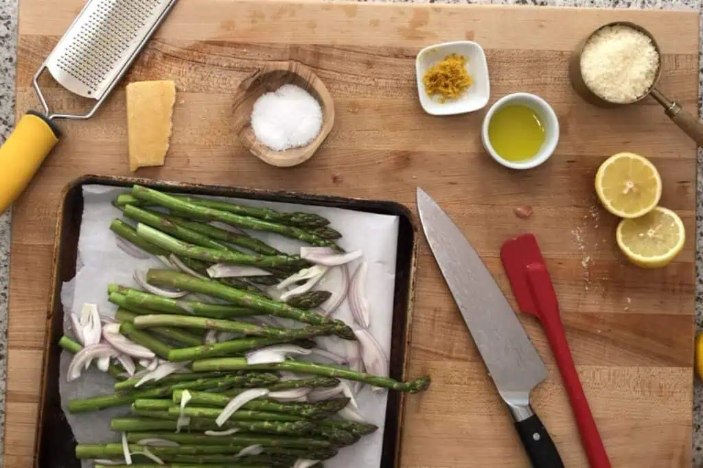 A cutting board with asparagus and sliced onions on a tray, a chef’s knife, lemon, grated cheese, olive oil, salt, lemon zest, a cheese grater, and a spatula arranged nearby.