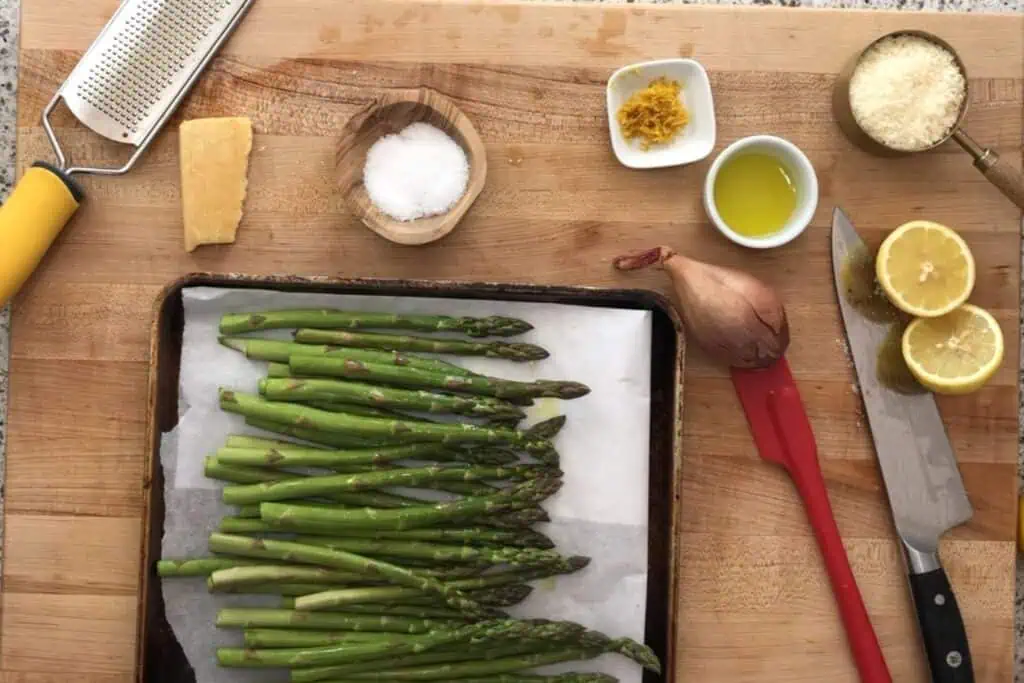 Overhead view of a tray of raw asparagus on parchment paper, surrounded by lemon halves, a shallot, olive oil, salt, parmesan, lemon zest, a grater, and a knife on a wooden surface.