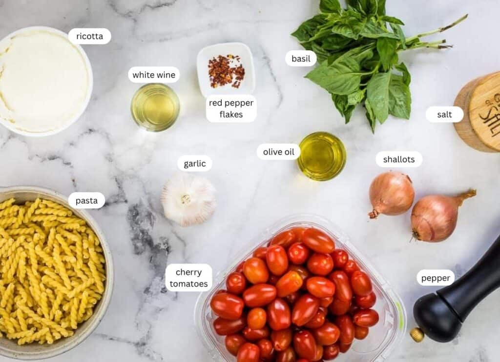 Ingredients for a pasta dish are arranged on a marble surface, including pasta, cherry tomatoes, ricotta, basil, shallots, garlic, olive oil, white wine, red pepper flakes, salt, and pepper.