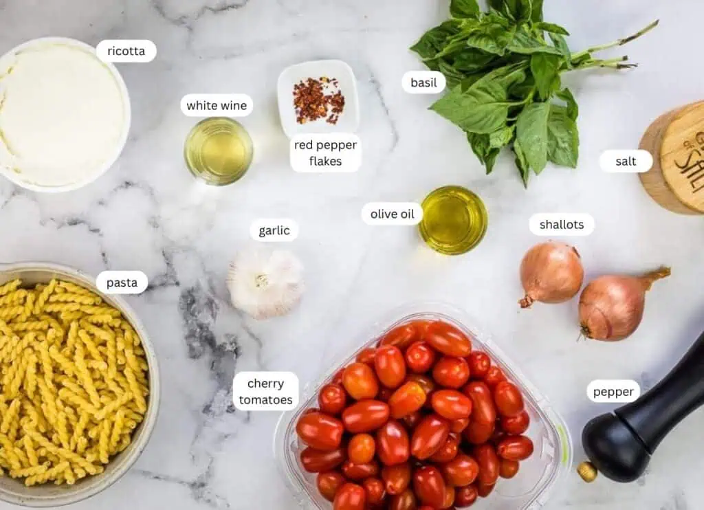 Ingredients for a pasta dish are arranged on a marble surface, including pasta, cherry tomatoes, ricotta, basil, shallots, garlic, olive oil, white wine, red pepper flakes, salt, and pepper.