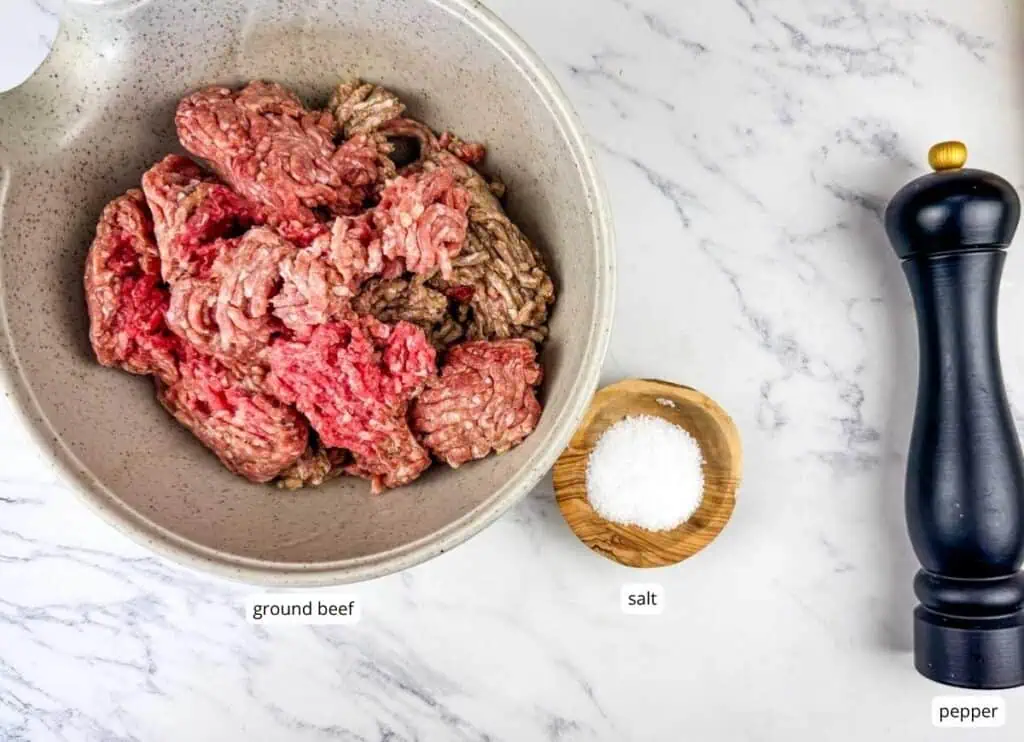 A bowl of ground beef, a small wooden bowl of salt, and a black pepper grinder on a marble countertop.