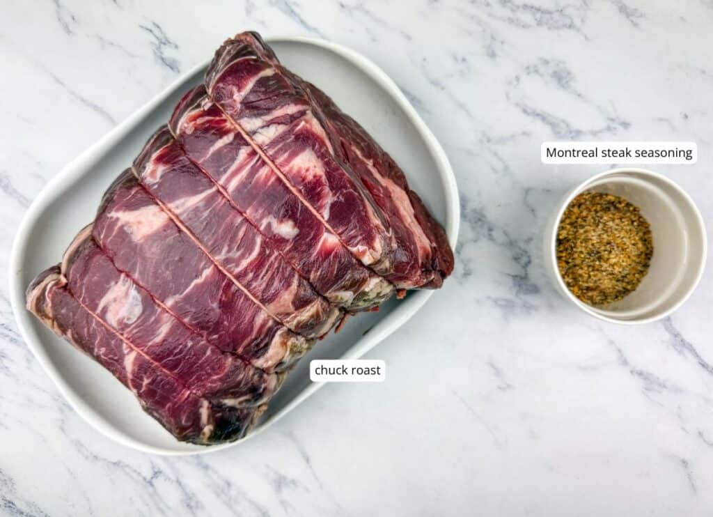 A raw chuck roast is tied with string on a white plate, next to a small bowl of Montreal steak seasoning on a marble surface. Labels identify each item.