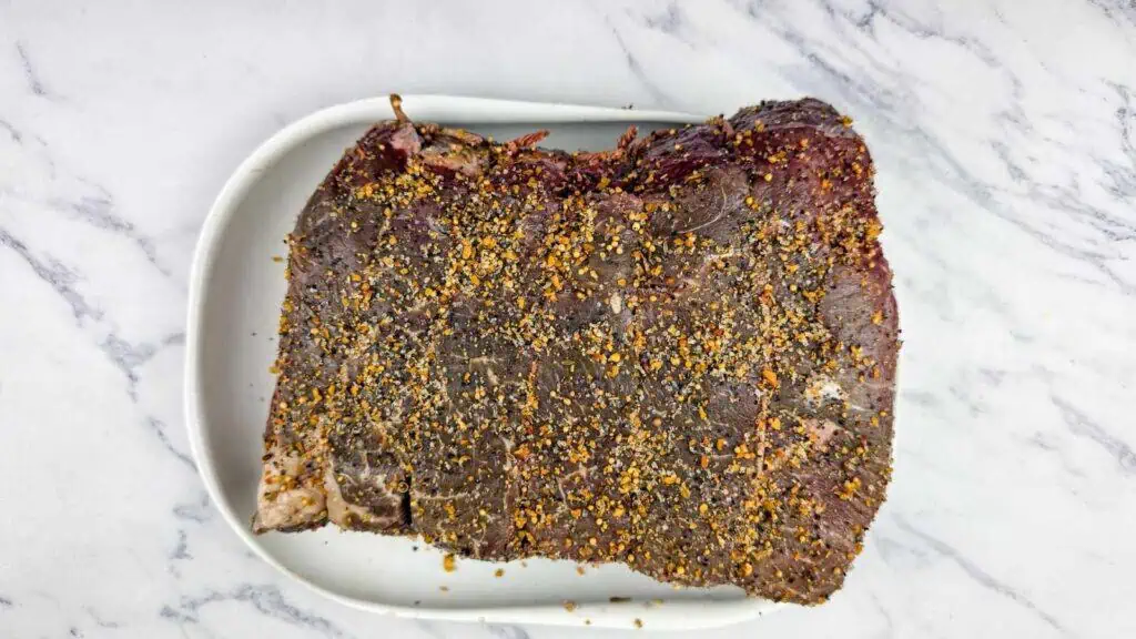 A large slab of seasoned raw beef sits on a white oval plate atop a marble surface.