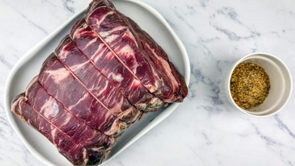 Raw tied beef roast on a white tray next to a small bowl of mixed spices on a marble surface.