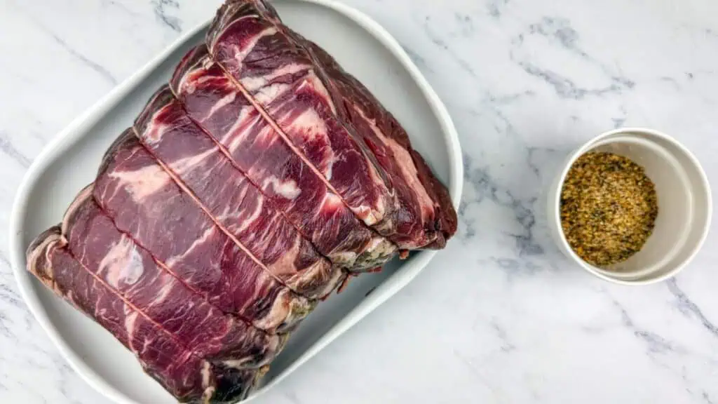 Raw tied beef roast on a white tray next to a small bowl of mixed spices on a marble surface.