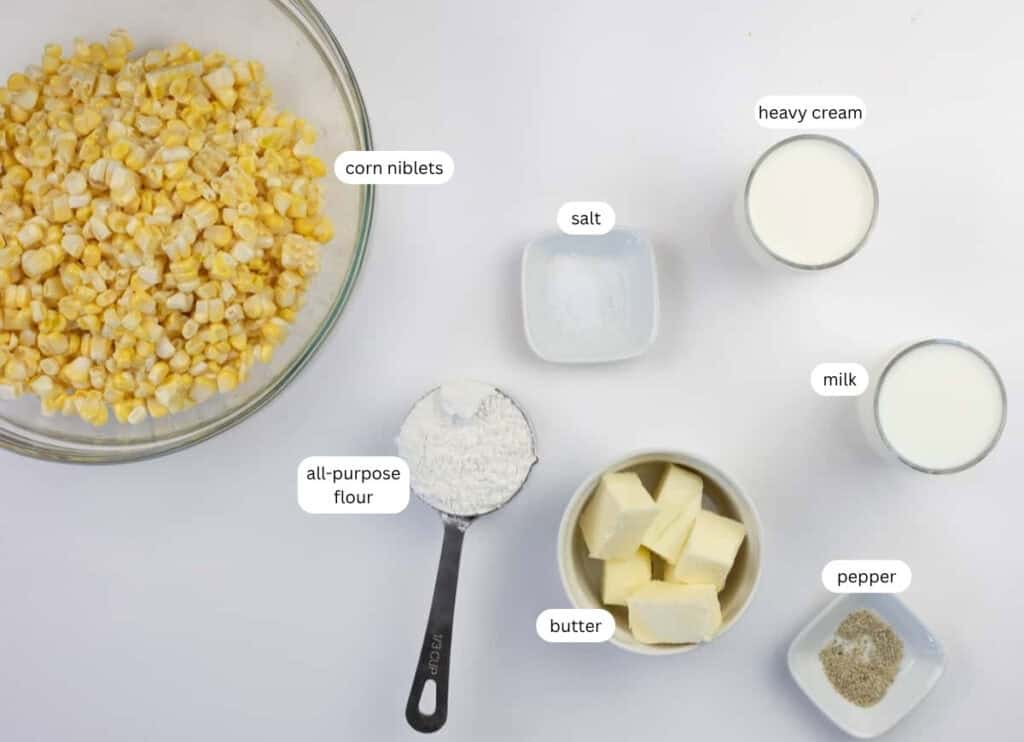 A bowl of corn niblets, a measuring cup of flour, a bowl of butter, and small bowls of salt, pepper, heavy cream, and milk arranged on a white surface.