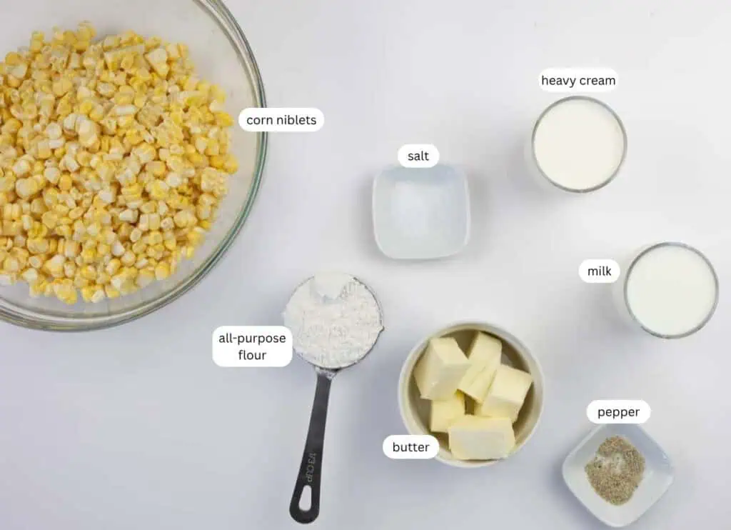 A bowl of corn niblets, a measuring cup of flour, a bowl of butter, and small bowls of salt, pepper, heavy cream, and milk arranged on a white surface.