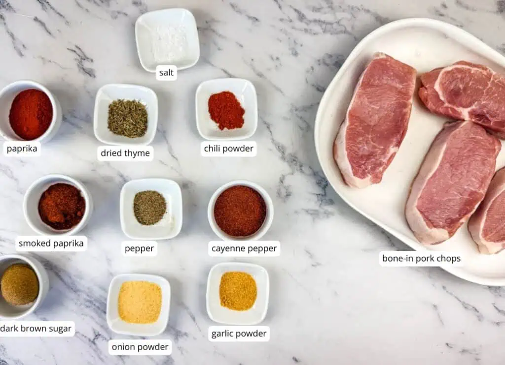 Ingredients for pork chops are arranged on a marble surface, including seasonings in small bowls and three raw bone-in pork chops on a white plate.