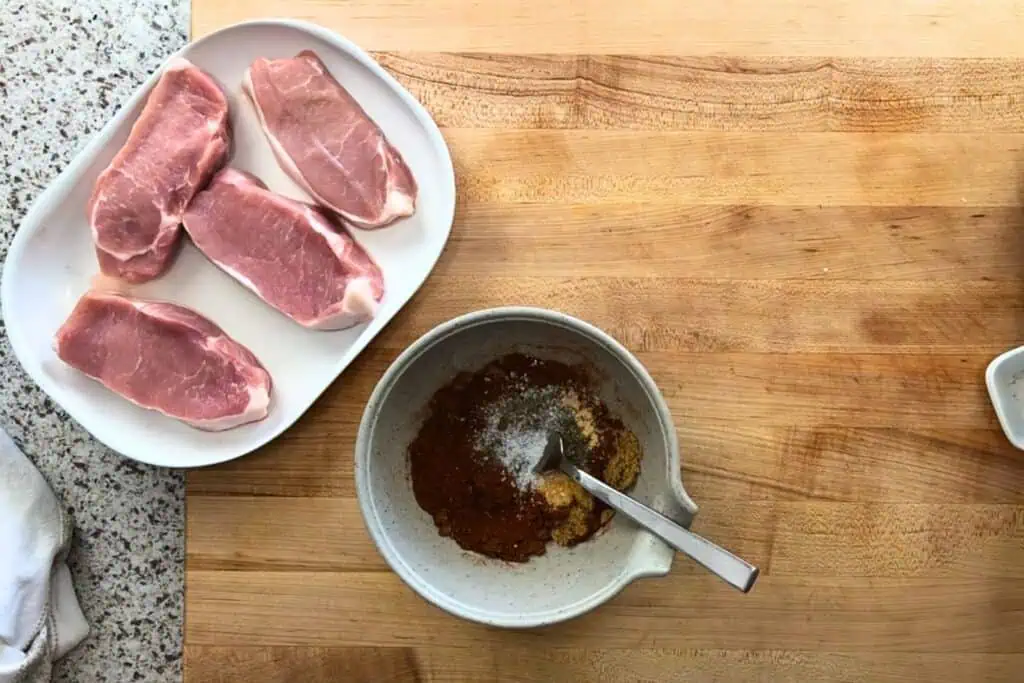 Four raw pork chops on a white plate next to a bowl containing mixed spices and a spoon, placed on a wooden countertop.