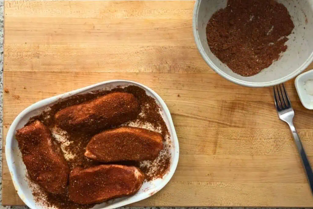 Four raw pork chops coated with dry rub seasoning in a white dish, next to a bowl of mixed spices, on a wooden cutting board with a fork nearby.