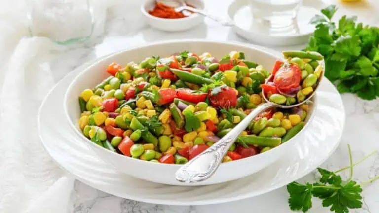 A white bowl filled with a colorful salad of corn, edamame, green beans, tomatoes, and parsley sits on a white plate with a silver spoon.