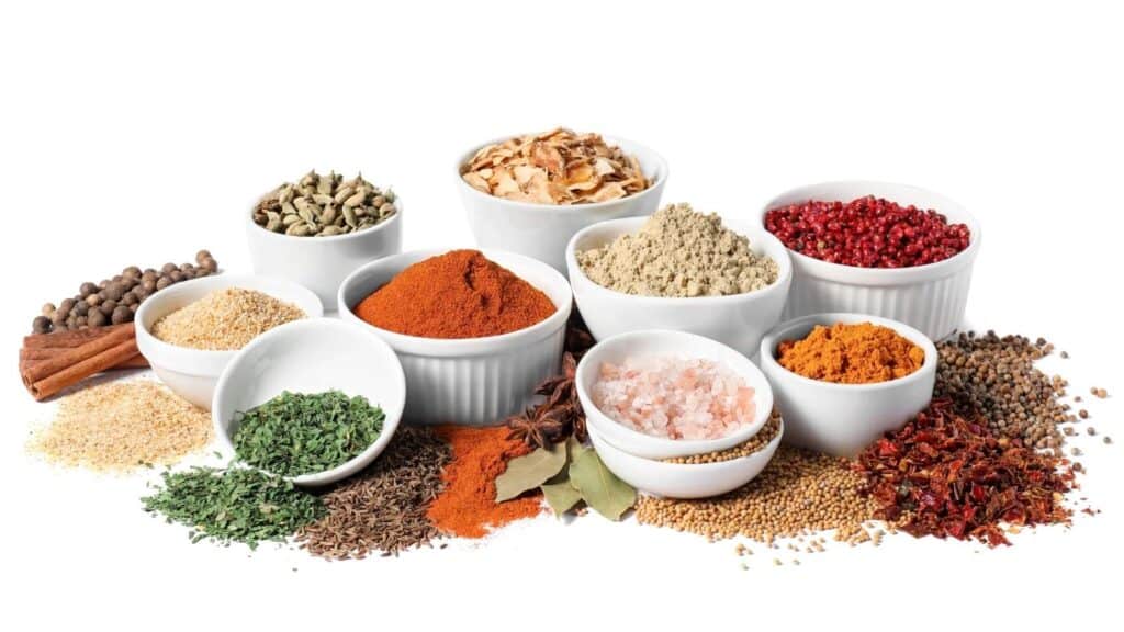 A variety of spices and herbs arranged in small white bowls and scattered on a white surface.