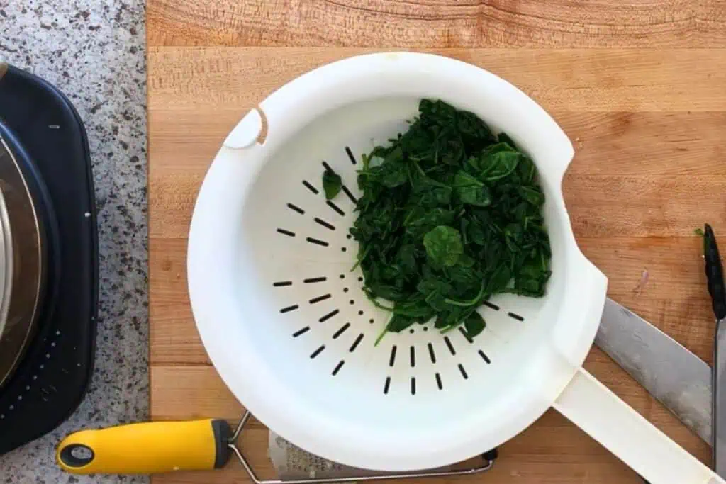 A white colander with cooked spinach sits on a wooden cutting board next to a knife and tongs.