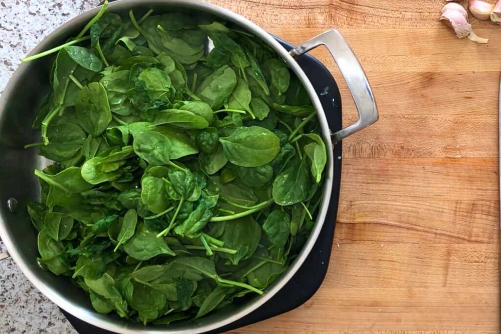 A large pot filled with fresh spinach leaves sits on a wooden cutting board, with garlic cloves visible in the top right corner.