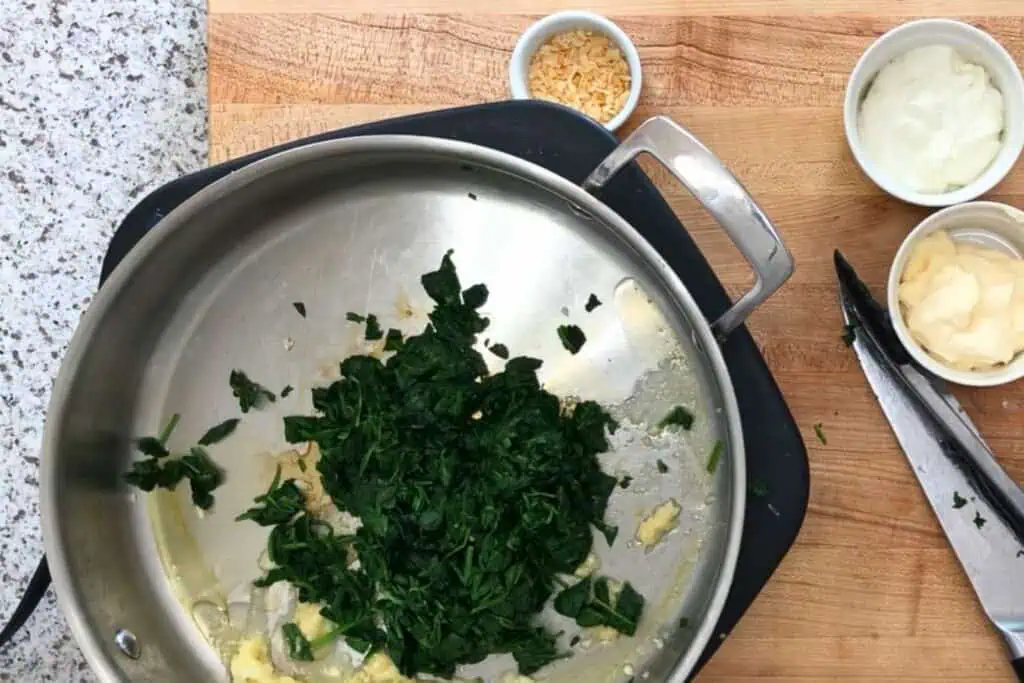 Chopped cooked spinach in a skillet on a wooden board, with bowls of minced garlic, mayonnaise, and sour cream nearby.