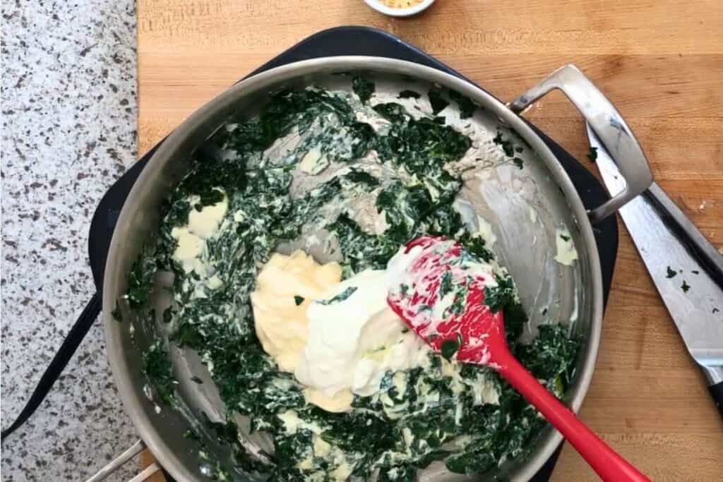 A red spatula mixes spinach, cream, and mayonnaise in a metal pan on a wooden countertop, with a knife and small bowl nearby.