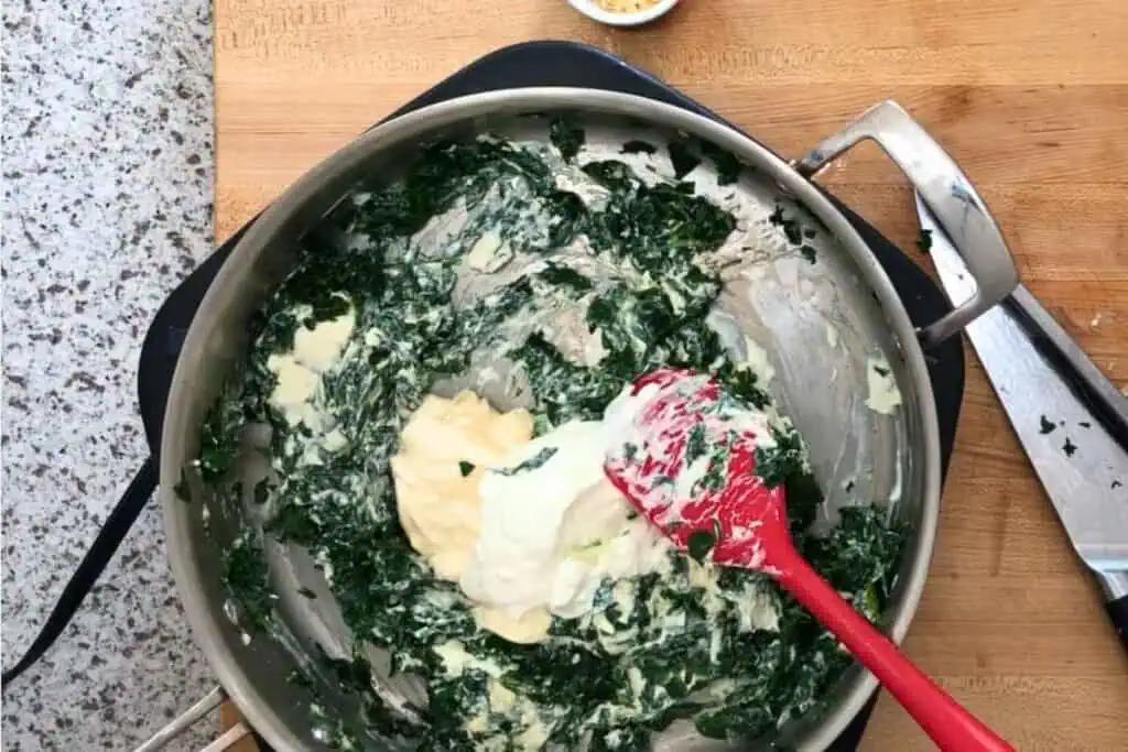 A red spatula mixes spinach, cream, and mayonnaise in a metal pan on a wooden countertop, with a knife and small bowl nearby.