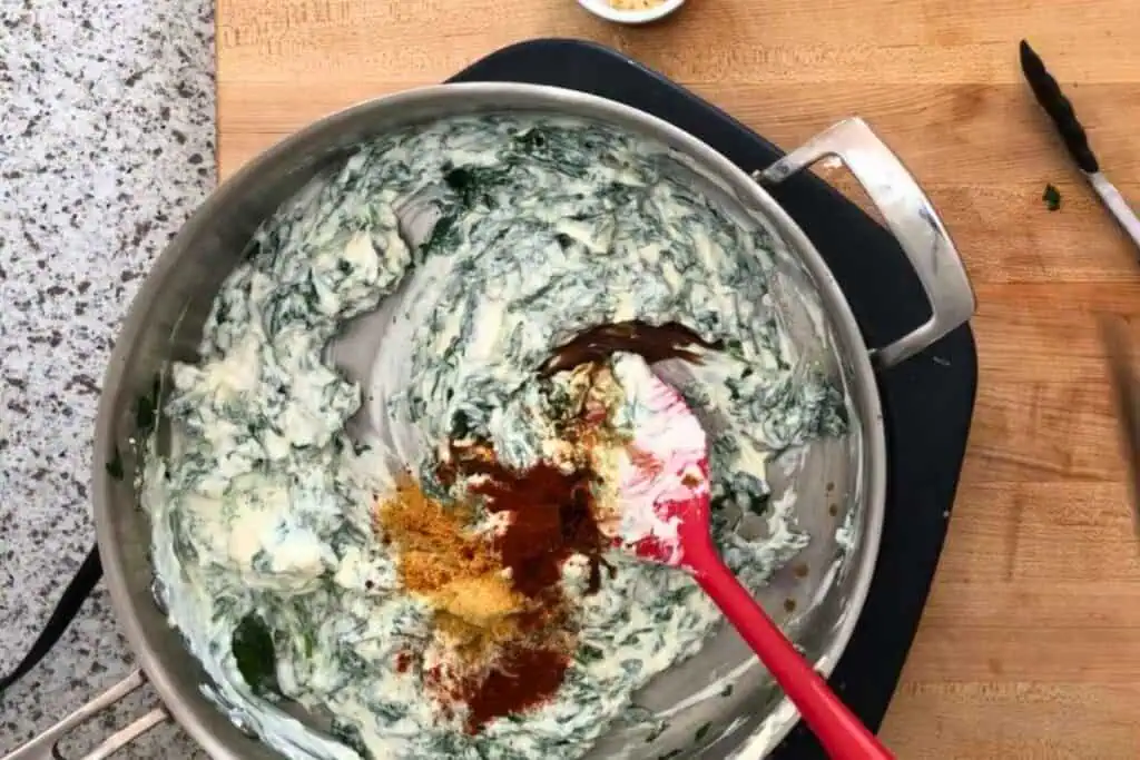 A metal pan on a stove contains creamy spinach dip being mixed, with spices including paprika, turmeric, and cumin added on top. A red spatula is in the pan, and tongs are nearby.