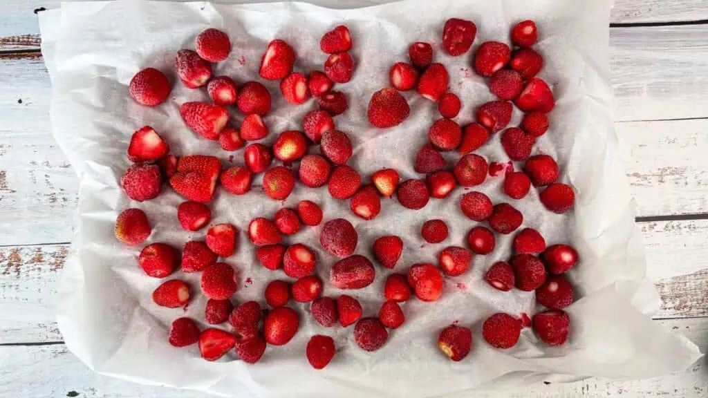 A tray lined with parchment paper holds an even layer of frozen strawberries spread out across the surface.