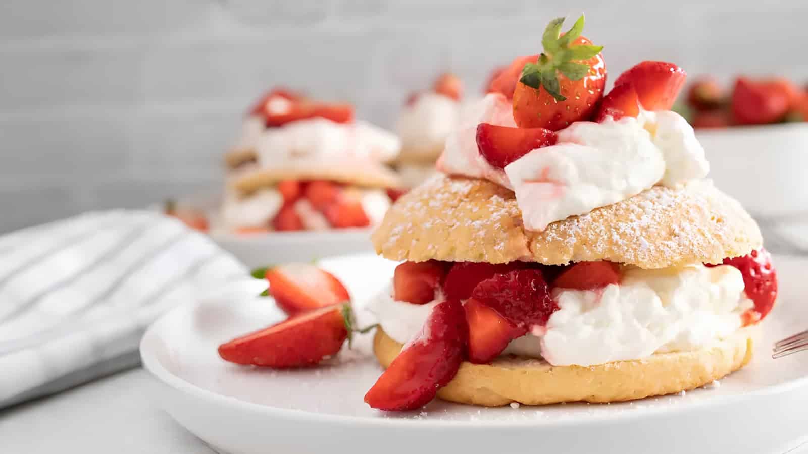 A strawberry shortcake with whipped cream and fresh strawberries is served on a white plate, with another dessert visible in the background.