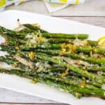 A white plate with roasted asparagus topped with grated cheese and lemon zest, garnished with lemon slices.
