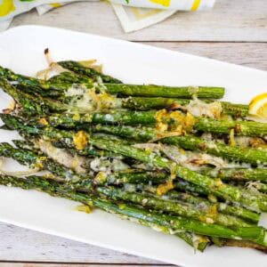 A white plate with roasted asparagus topped with grated cheese and lemon zest, garnished with lemon slices.