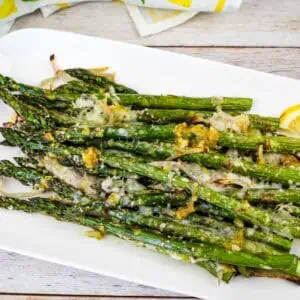 A white plate with roasted asparagus topped with grated cheese and lemon zest, garnished with lemon slices.