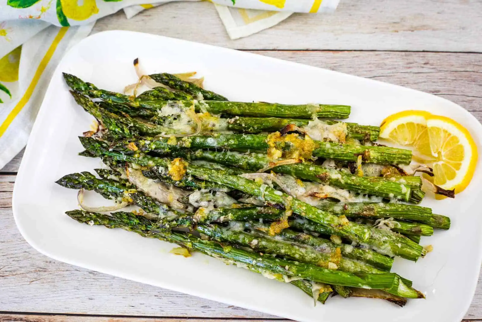 A white plate with roasted asparagus topped with grated cheese and lemon zest, garnished with lemon slices.