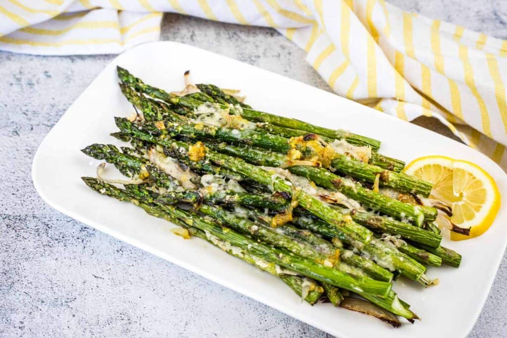 A plate of roasted asparagus topped with melted cheese, garnished with lemon slices, on a white rectangular dish with a yellow-striped cloth nearby.