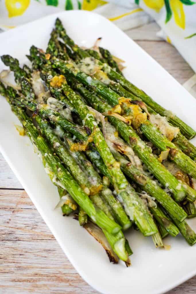Roasted asparagus spears topped with melted cheese and seasoning are arranged on a white rectangular plate.