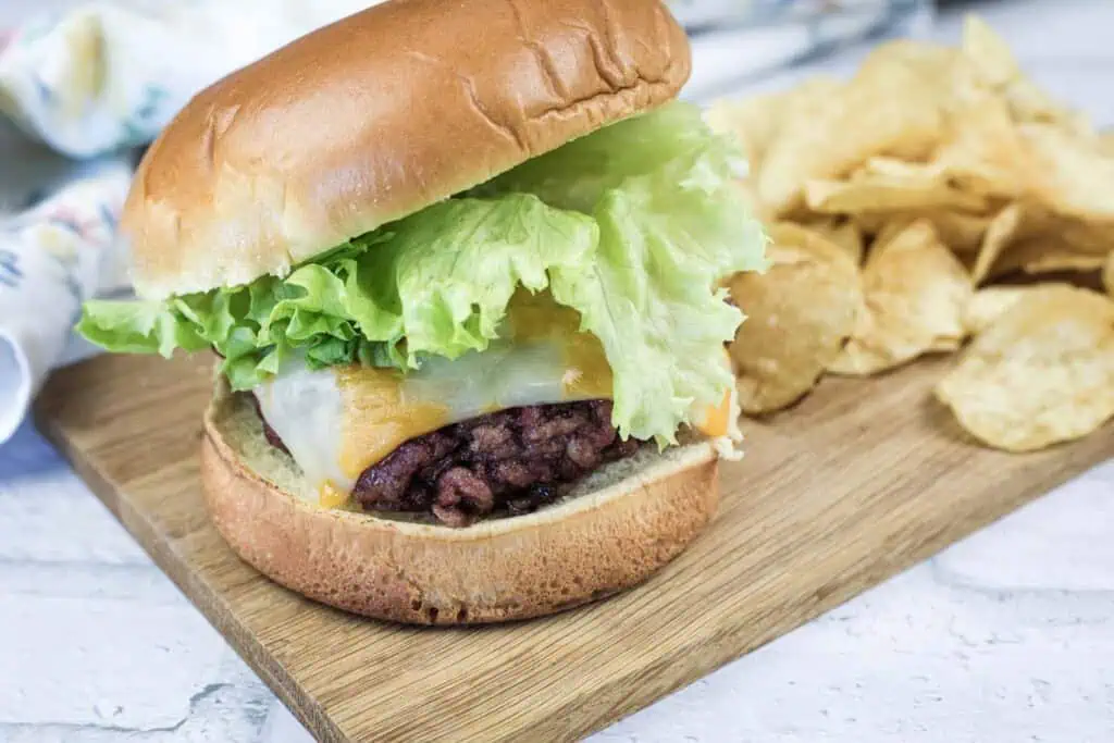 A hamburger with lettuce, cheddar and Swiss cheese on a bun is served on a wooden board next to a pile of potato chips.