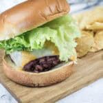 A hamburger with lettuce, cheddar and Swiss cheese on a bun is served on a wooden board next to a pile of potato chips.