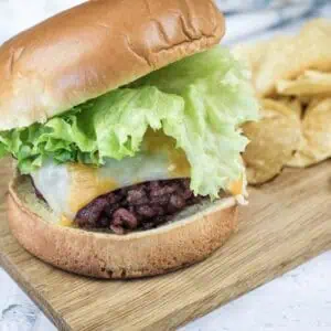 A hamburger with lettuce, cheddar and Swiss cheese on a bun is served on a wooden board next to a pile of potato chips.