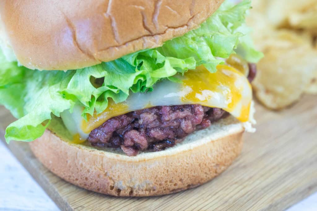 A close-up of a hamburger with lettuce, melted cheese, and a beef patty on a bun, served on a wooden surface with potato chips in the background.