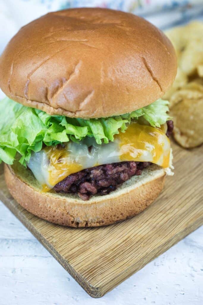 A cheeseburger with lettuce, melted cheese, and a beef patty on a bun, served on a wooden board next to potato chips.