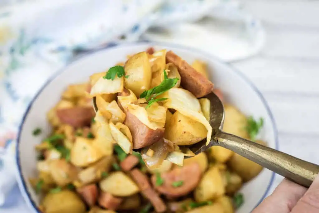 A spoonful of cooked potatoes, sausage, and cabbage held above a bowl filled with the same mixture, garnished with parsley.