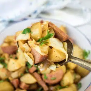 A spoonful of cooked potatoes, sausage, and cabbage held above a bowl filled with the same mixture, garnished with parsley.