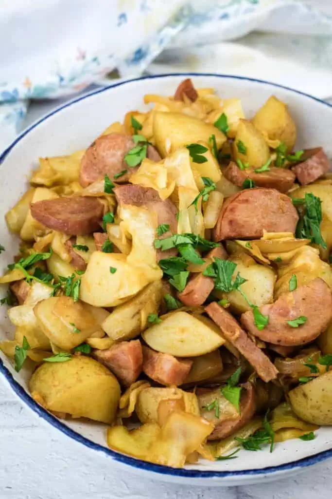 A bowl of sautéed potatoes, sausage, and cabbage garnished with chopped parsley, served on a white plate.