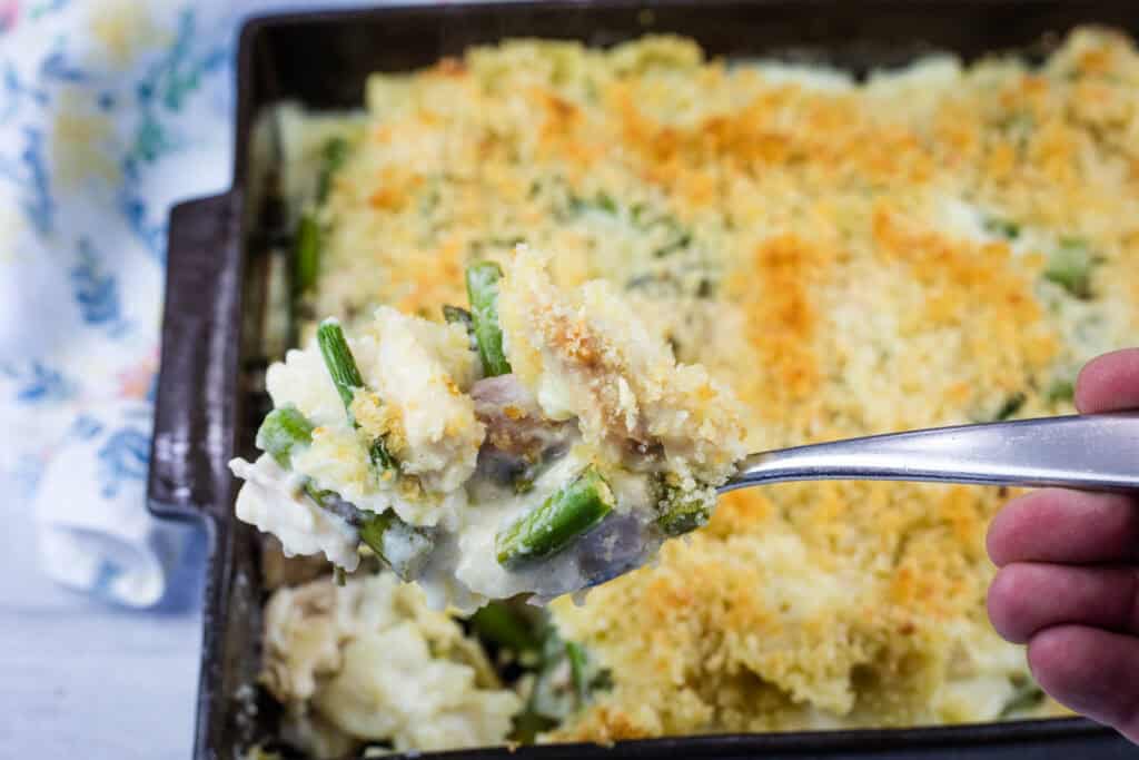A hand holds a spoonful of creamy vegetable casserole with a breadcrumb topping above a baking dish filled with the same casserole.