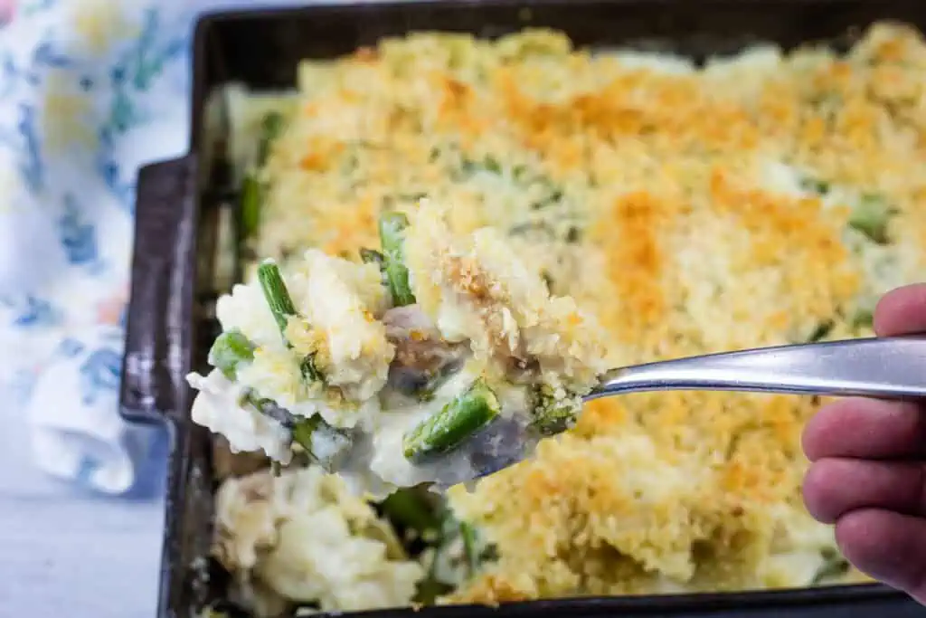 A hand holds a spoonful of creamy vegetable casserole with a breadcrumb topping above a baking dish filled with the same casserole.