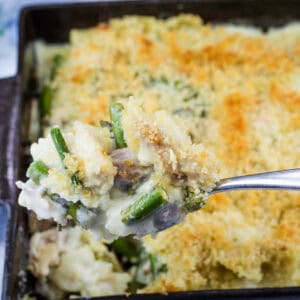 A hand holds a spoonful of creamy vegetable casserole with a breadcrumb topping above a baking dish filled with the same casserole.